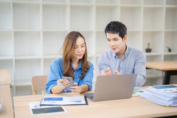 Two asian businesspeople are working together in a modern office, using laptop and smartphone to...