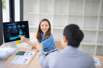 Two business professionals discussing data analytics displayed on a computer screen, focusing on graphs and charts, in a modern office setting with shelves in the background