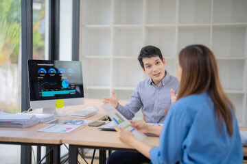Two colleagues engage in a discussion, analyzing data displayed on a computer screen. Papers and graphs are spread across the desk, indicating a focus on business strategy and planning
