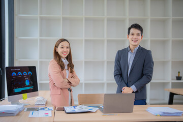 Two asian businesspeople standing behind a desk with arms crossed, smiling and posing in a modern office, with documents and laptop on the desk