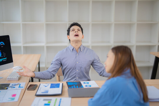 Stressed asian businessman complaining to his colleague about bad financial results, sitting at desk in office with computer and documents with charts and graphs