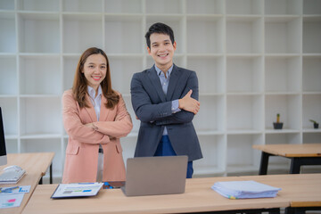 Two Asian businesspeople standing confidently with arms crossed in a modern office, smiling beside a desk with a laptop and paperwork, embodying teamwork and success