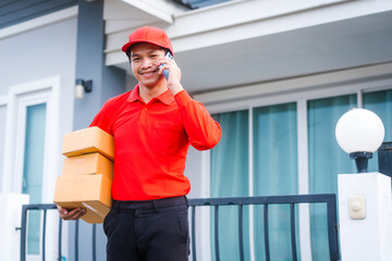 A cheerful delivery man in a red uniform and cap holds stacked packages while talking on his smartphone outside a modern, light-colored house.