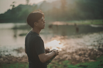 A young man stands by a tranquil lake at sunset, gazing thoughtfully into the distance with soft sunlight reflecting on the water.
