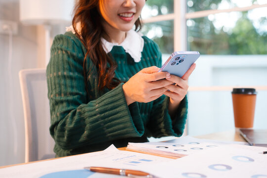 Confident middle-aged Asian professional works on a laptop amidst scattered charts and documents in a bright home office, a coffee cup nearby hinting at a dedicated work session.