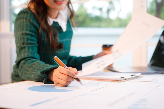 Confident middle-aged Asian professional works on a laptop amidst scattered charts and documents in a bright home office, a coffee cup nearby hinting at a dedicated work session.
