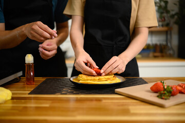 Close up of couple decorating pancakes with fresh strawberries in a kitchen