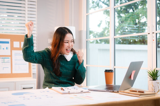 Confident middle-aged Asian professional works on a laptop amidst scattered charts and documents in a bright home office, a coffee cup nearby hinting at a dedicated work session.