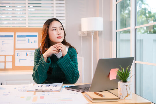 Confident middle-aged Asian professional works on a laptop amidst scattered charts and documents in a bright home office, a coffee cup nearby hinting at a dedicated work session.