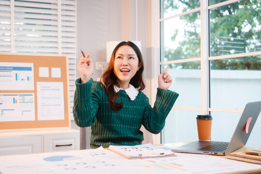 Confident middle-aged Asian professional works on a laptop amidst scattered charts and documents in a bright home office, a coffee cup nearby hinting at a dedicated work session.