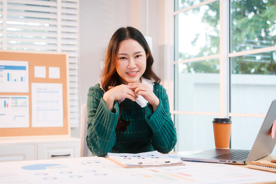 Confident middle-aged Asian professional works on a laptop amidst scattered charts and documents in a bright home office, a coffee cup nearby hinting at a dedicated work session.
