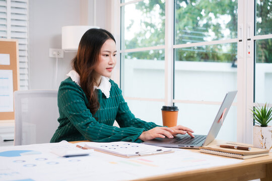 Confident middle-aged Asian professional works on a laptop amidst scattered charts and documents in a bright home office, a coffee cup nearby hinting at a dedicated work session.
