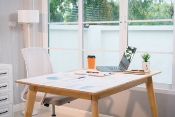 A bright, modern office space with a wooden desk covered in documents, charts, a laptop, and a...