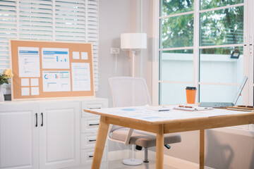 A bright, modern office space with a wooden desk covered in documents, charts, a laptop, and a coffee cup. A white chair and large windows create a clean, productive atmosphere.