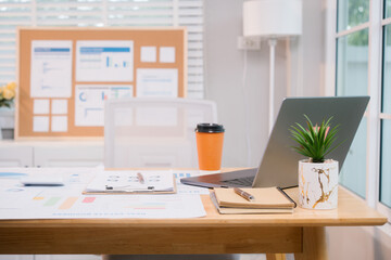 A bright, modern office space with a wooden desk covered in documents, charts, a laptop, and a...