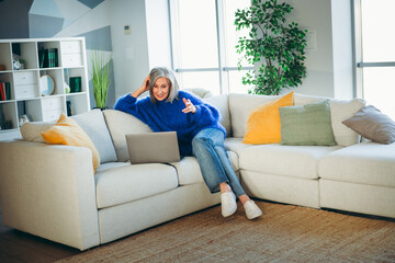 Obraz premium Mature woman relaxing on a sofa with a laptop, enjoying leisure time and comfort in a stylish indoor living room