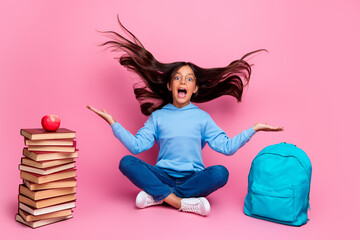 Excited young girl smiling with flying hair sitting near book and backpack in a pink studio background