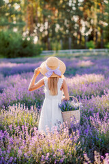 girl in a hat stands in a field of lavender and looks at the sunset. She's wearing a white dress. There's a basket of lavender in her hand. View from the back