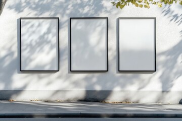 Three blank, vertical, framed posters mounted on a white wall outdoors, casting shadows from an overhanging tree's leaves onto the wall and sidewalk