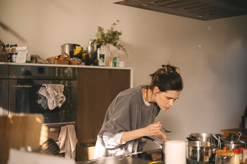 Woman standing by the stove in the kitchen, cooking and smelling the nice aromas from her meal in a...