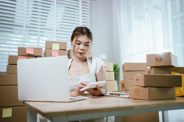 Young Asian businesswoman packing online orders at home office, using laptop for e-commerce sales and shipping in a small business.