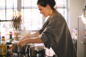Young woman cooking in a cozy kitchen, focused on her task while opening a hot pot on the stove. Natural light, casual clothes, warm home atmosphere, real moment from everyday domestic life.