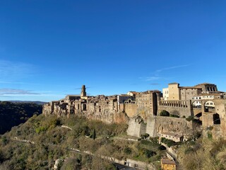 Fototapeta premium Pitigliano medieval village clinging to tufa cliff, showcasing historic stone dwellings against vibrant blue sky in scenic Tuscan landscape