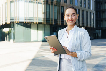 Businesswoman in modern city smiling while holding tablet during bright sunny day