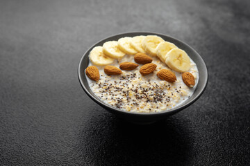 Oatmeal porridge with banana slices almonds and chia seeds in dark bowl on black background