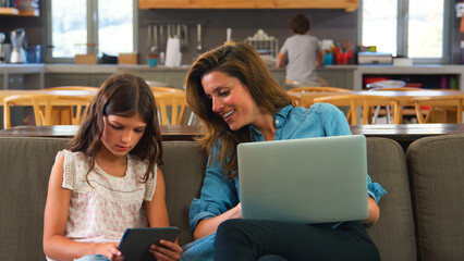Mother And Daughter Sitting On Lounge Sofa Using Digital Tablet And Laptop