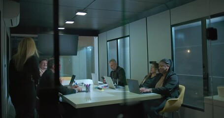 Female executive walking into boardroom to address coworkers during business meeting with diverse team in modern office at night - Powered by Adobe