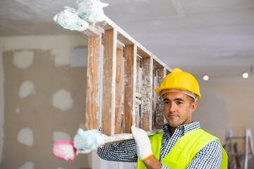 Young foreman in uniform working on a construction site indoors during the repair carries a stepladder from one room to another