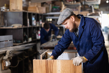 Young male carpenter assembles wooden cabinet from parts in wood workshop