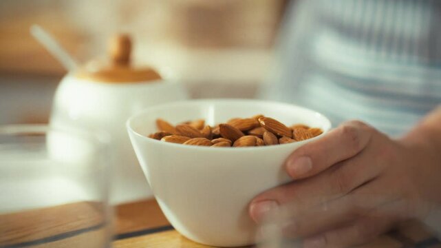 A person gently takes almonds from a white bowl while preparing a snack in a well-lit kitchen. The setting creates a warm and inviting atmosphere, showcasing a moment of mindful eating
