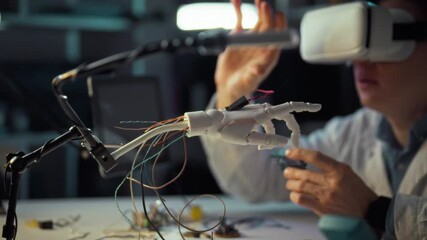 An engineer conducts an experiment using a virtual reality headset while manipulating a robotic hand. This action takes place in a lab setting filled with technology