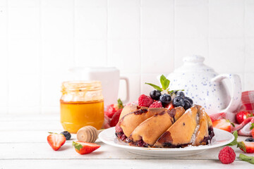 Sweet summer bundt cake with fresh fruit and berry. Traditional round sponge cake, seasonal dessert with strawberry, raspberry, blueberry, honey and sugar sprinkles, white wooden table copy space