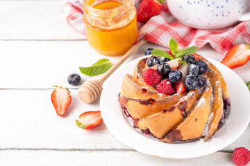 Sweet summer bundt cake with fresh fruit and berry. Traditional round sponge cake, seasonal dessert with strawberry, raspberry, blueberry, honey and sugar sprinkles, white wooden table copy space