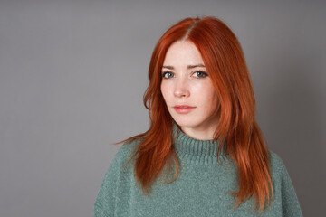 Portrait of young Ukrainian woman with dyed red hair and freckles wearing green sweater on gray background with copy space