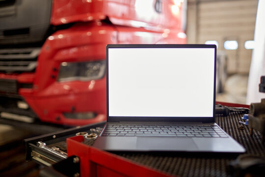 Open laptop placed on mechanic's workbench with tools nearby and large red truck in background, suggesting an automotive repair setting