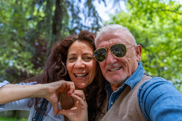 beautiful middle aged couple taking selfie showing heart symbol with hand outdoors