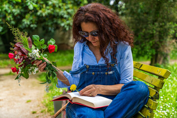 middle-aged woman with red hair sitting on a bench reading a book holding a bouquet of flowers
