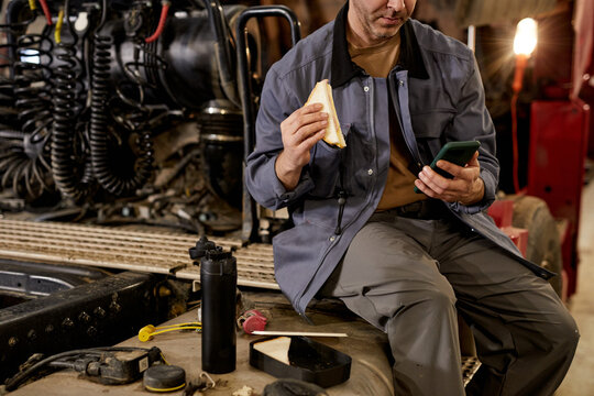 Mechanic sitting in workshop taking break by eating sandwich and using phone. Surrounded by various tools and equipment in industrial setting