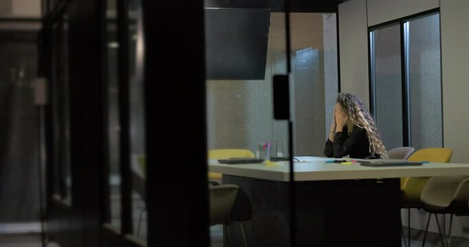 Young woman sits alone in glass-walled office with face buried in hands, exhausted and emotionally drained following high-pressure corporate workday