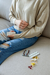 Young caucasian female experiencing stomach pain with various pills and blisters on couch