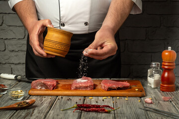A chef sprinkles salt over raw cuts of meat placed on a wooden cutting board. Various spices and...