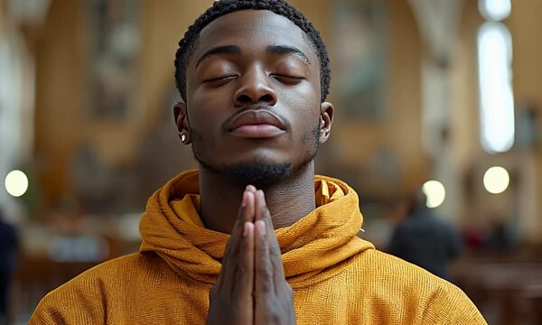 Young man with eyes closed, hands clasped in prayer, wearing a mustard yellow hoodie, in a blurred background.
