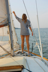 A girl in a striped shirt stands on the bow of a sailing yacht and waves her hand, back view.