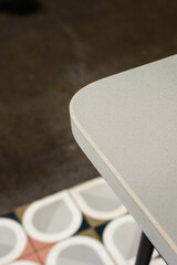 Smooth curve. Close-up of a light grey speckled surface with a rounded edge, contrasting with a blurred background of dark floor and patterned tiles.