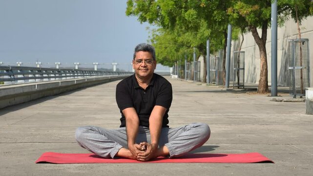 Senior Indian man practicing yoga outdoors in butterfly pose - Baddha Konasana on a red mat, enjoying a peaceful morning workout session by the riverside in Ahmedabad, India