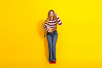 Happy young woman wearing striped sweater and jeans holding book on bright yellow background, looking cheerful